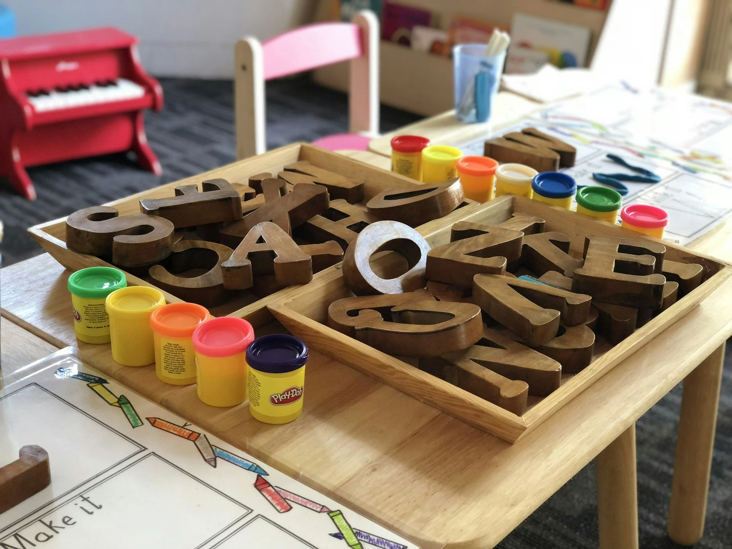 Wooden alphabet letters on a tray, colorful playdough containers, and kids' drawings on a table.