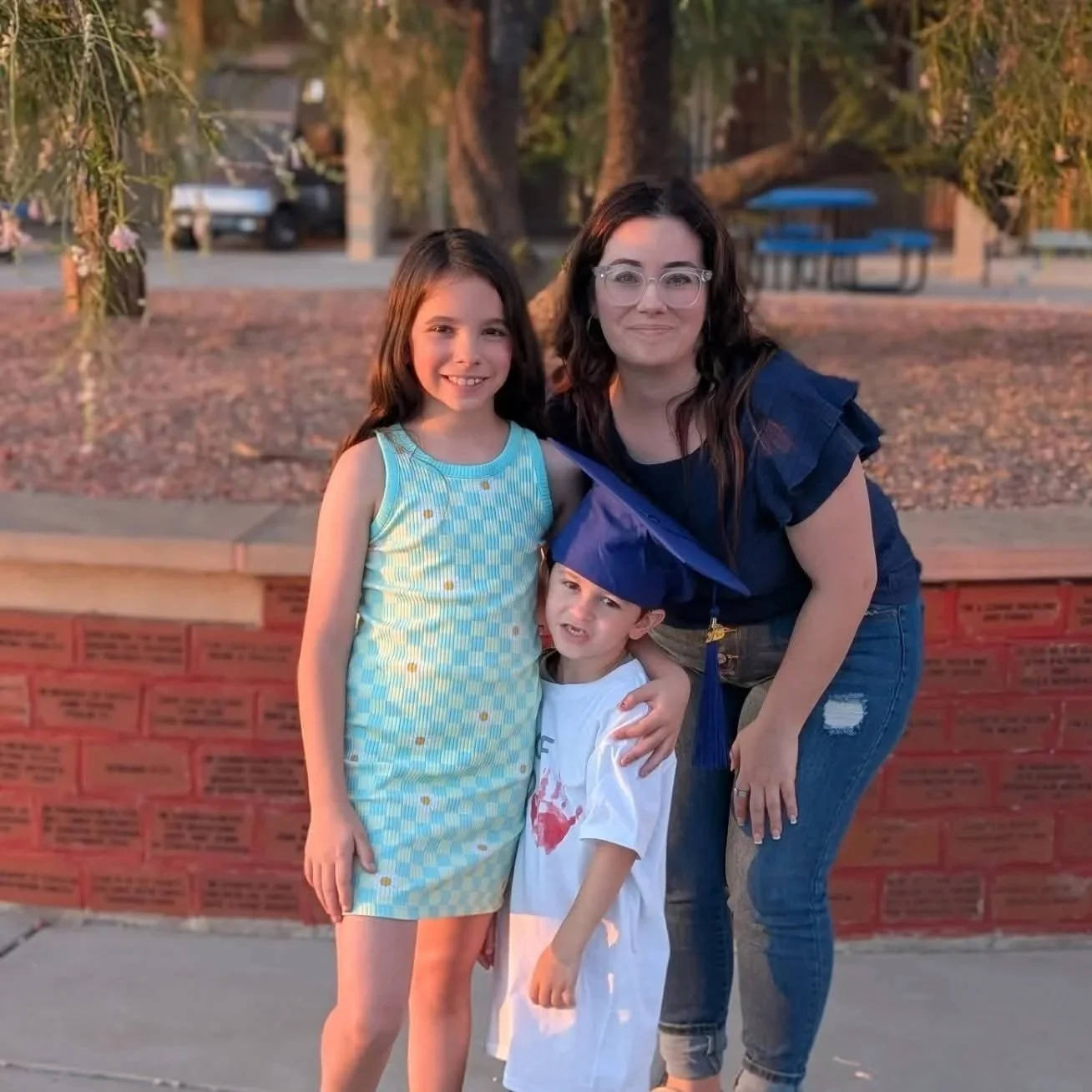 A woman with glasses and dark curly hair posing outdoors with two children, one girl and a young boy wearing a graduation cap, in front of a low red brick wall and a park in the background during sunset.