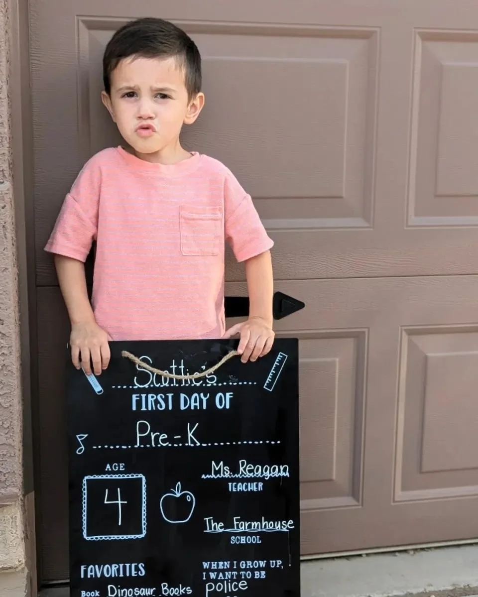 Young boy standing outside near a garage door, holding a black chalkboard sign with his first day of school information written on it, including his name, age, teacher, school, and favorite things.