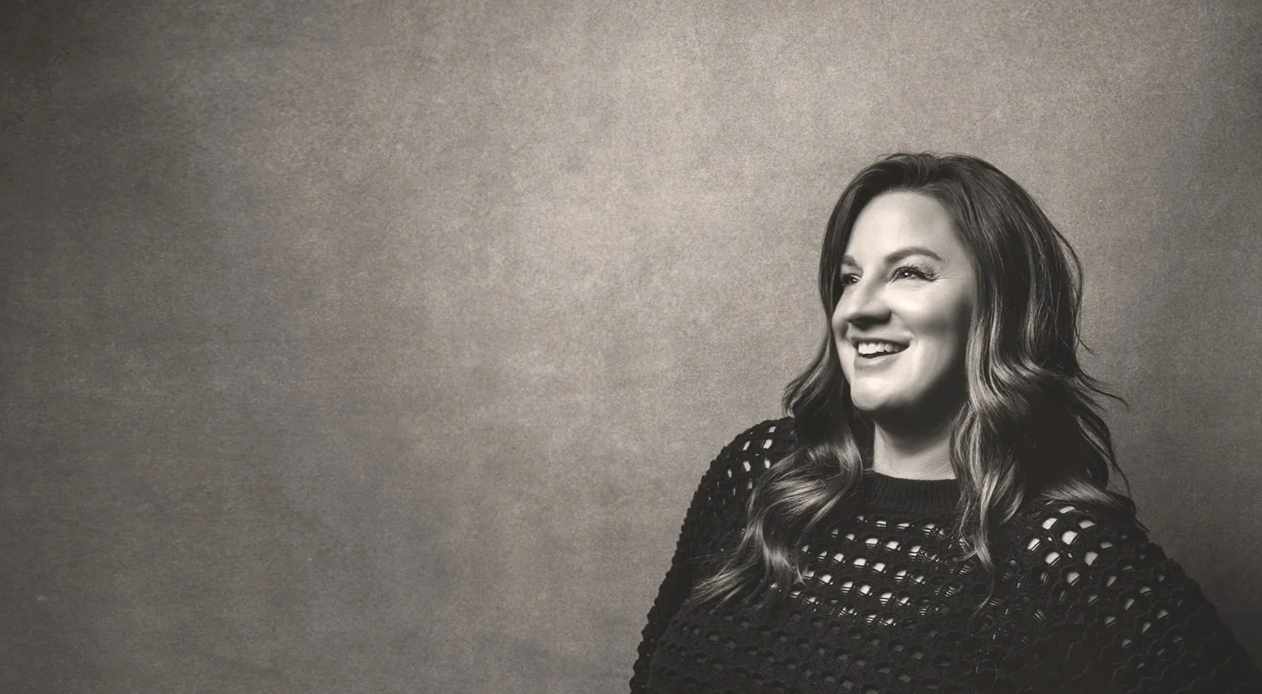 A woman with long, wavy hair smiling and looking to her left in a black, open-knit sweater. Black and white photo with a plain background.