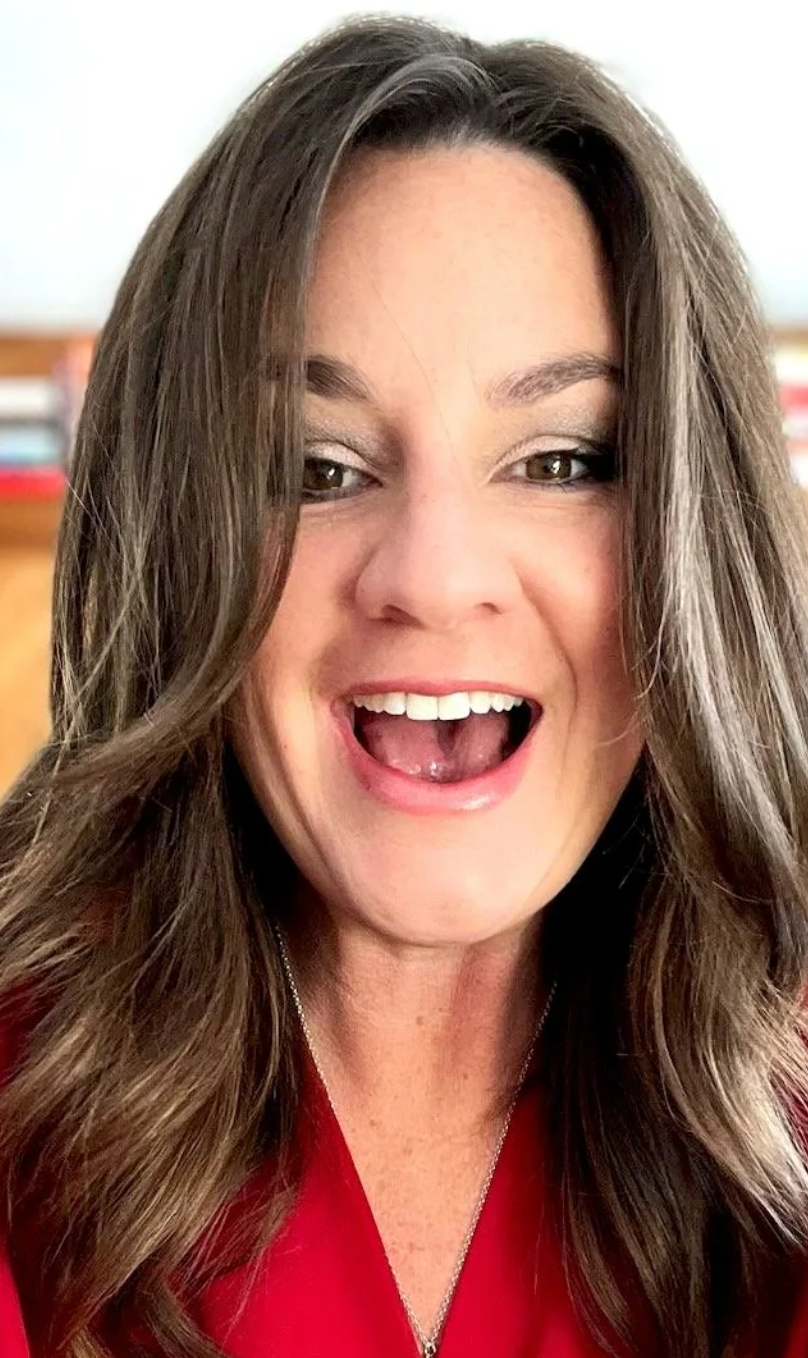 A woman with long brown hair, smiling with her mouth open, wearing makeup and a red top, indoors with blurred shelves in the background.