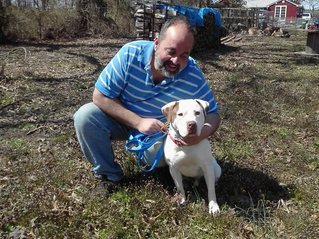 A man in a blue striped shirt kneeling on grass, holding a white dog with brown spots on a leash, outdoors on a sunny day.