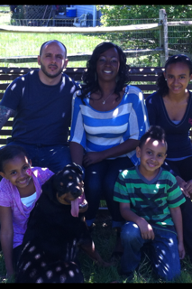 Family of five, including two children and a black dog, sitting on grass in park with a wooden fence and trees in background.