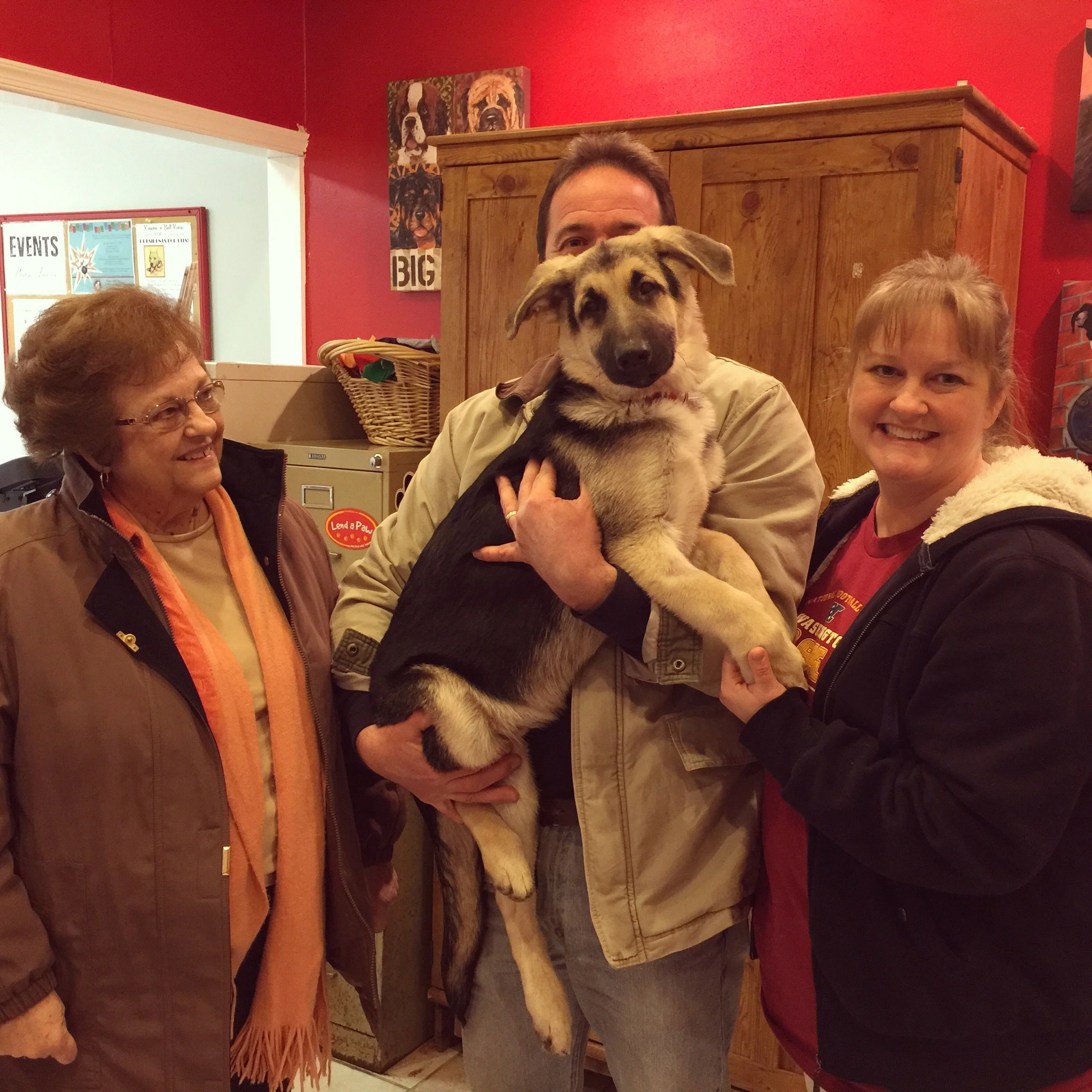 A man holding a puppy with a woman beside him, others in the background, in a room with red walls and wall art of dog portraits.