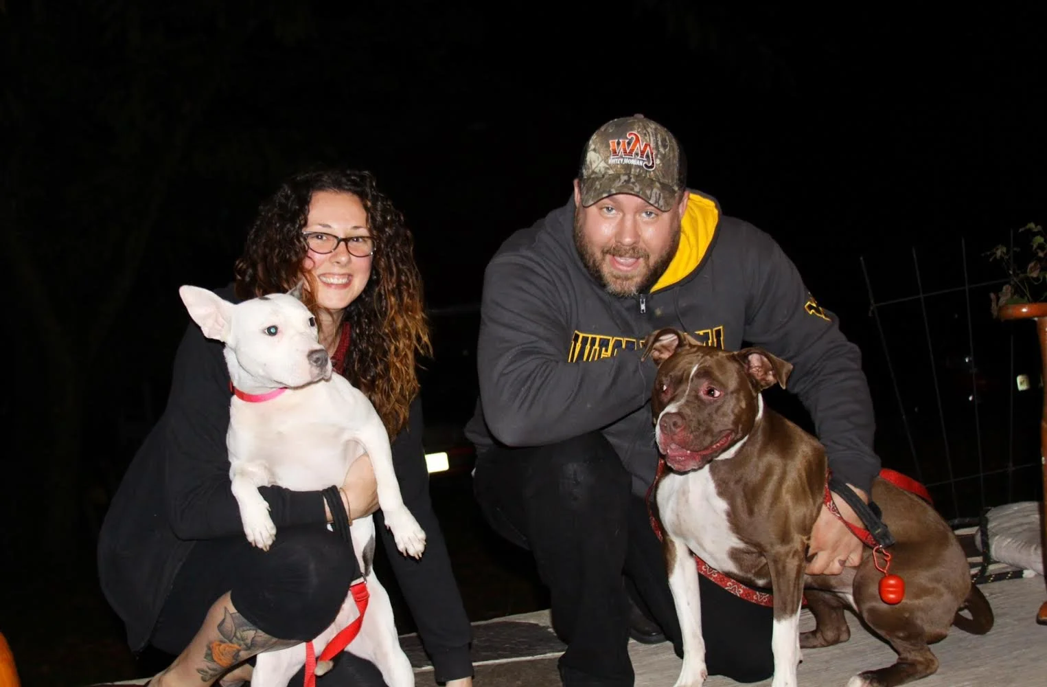 A woman with glasses and curly hair holding a white dog, and a man with a beard and wearing a camouflage cap with dogs, on a wooden deck outdoors at night.