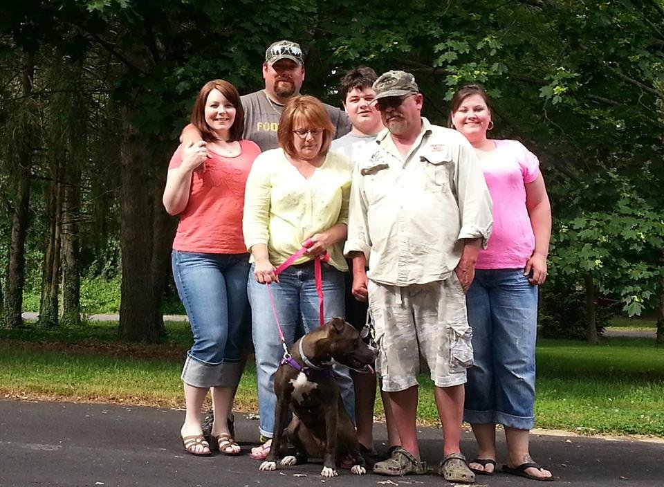 Group of six people and a dog standing outdoors on a paved path with trees in the background.