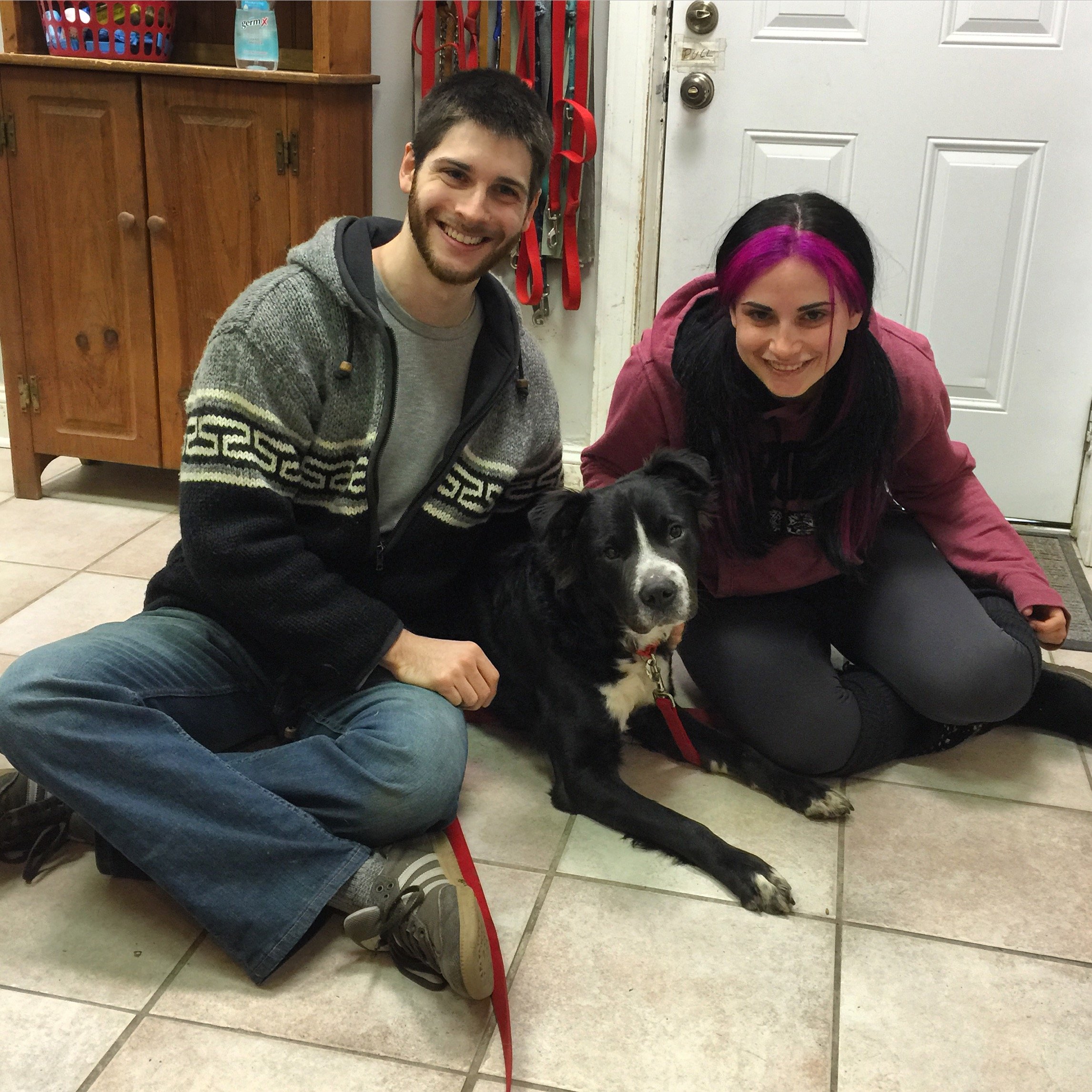 Two young adults, a man with short dark hair and beard and a woman with black hair dyed pink, are smiling and sitting on the floor next to a black and white dog. They are in a room with light-colored tiled floor, wooden cabinets, a white door with hi