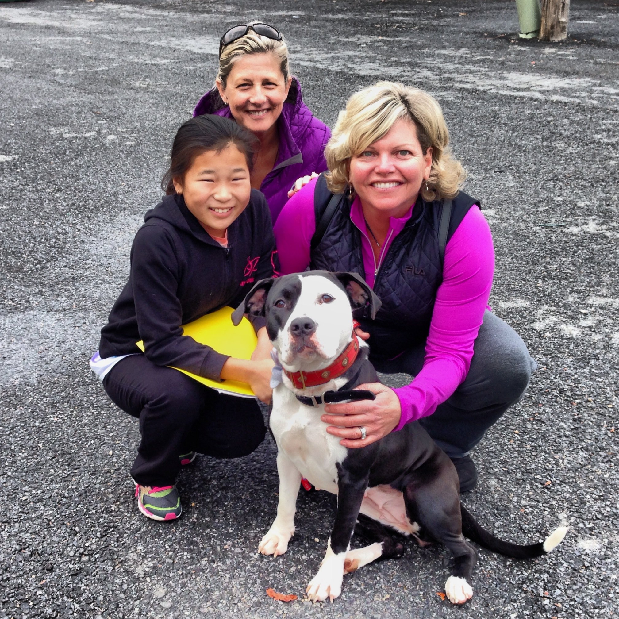 Three women and a young girl with their dog outside on a gravel surface, smiling and crouching together.