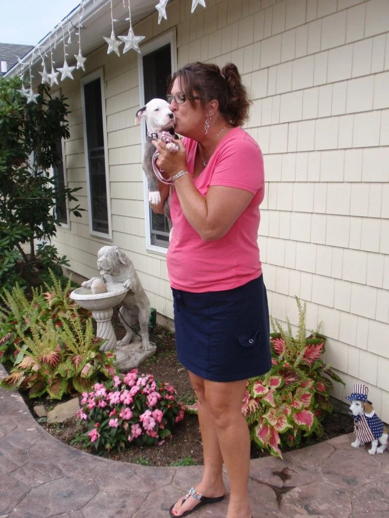 Woman in pink shirt and navy skirt holding and kissing a small white puppy outside a house decorated with star and American flag-themed decorations, with garden statues and plants nearby.