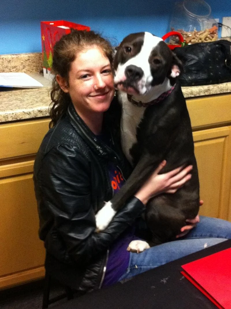 A woman with curly hair wearing a black leather jacket sits at a counter and hugs a large black and white dog, possibly a pitbull mix, in a kitchen.