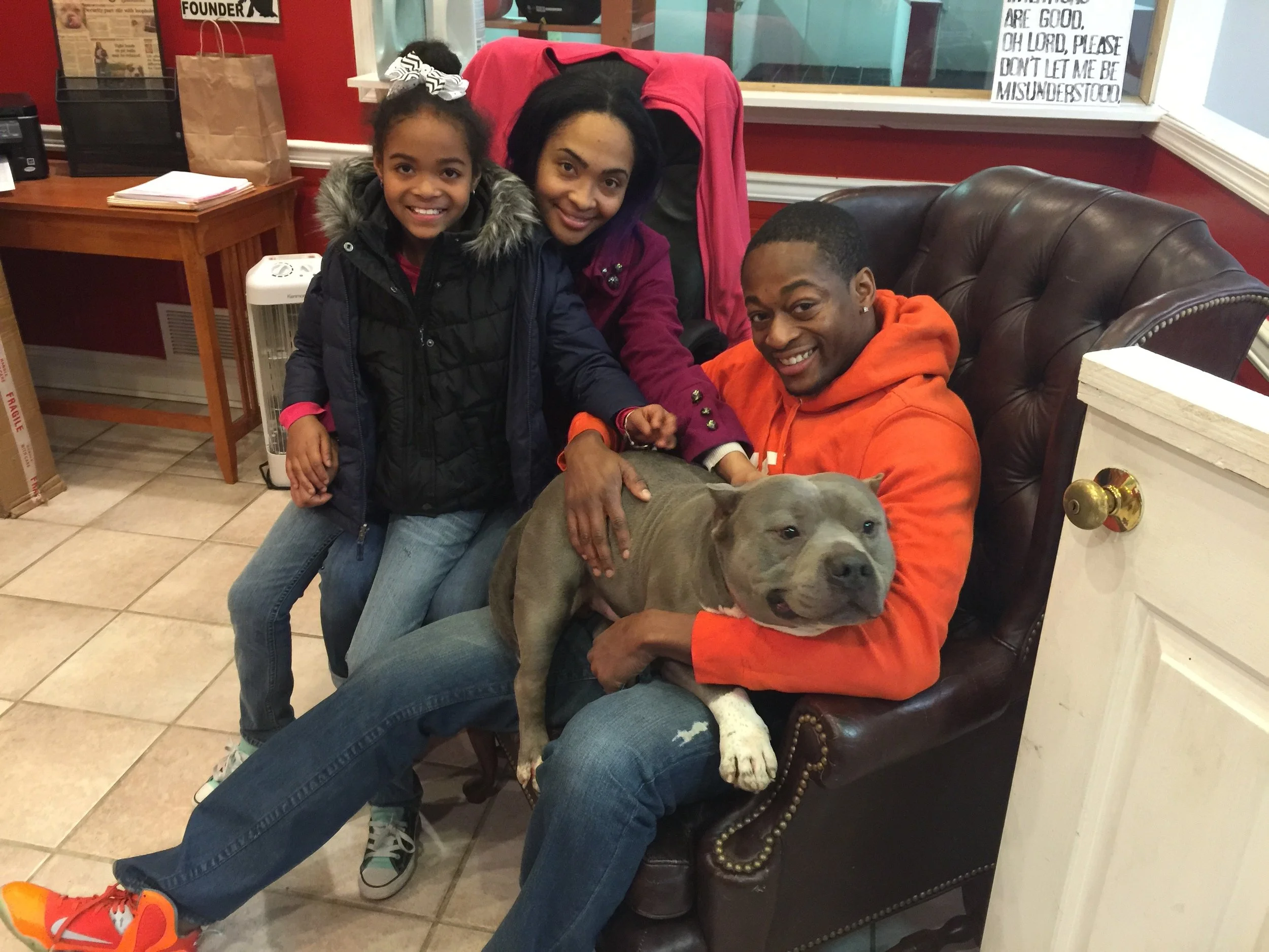 A woman and a girl sitting on a leather armchair with a gray dog in the foreground. The woman is wearing an orange hoodie, and the girl is wearing a black jacket with a fur-lined hood and a large white bow in her hair. Inside the room, there is a woo