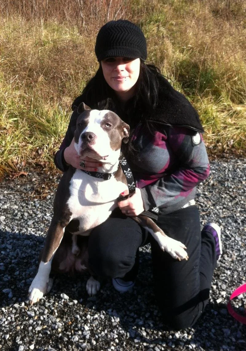 A woman in a black beanie and dark outdoor clothing kneels on a gravel path, holding a large, brown and white dog with a black collar. The background features tall, dry grasses.