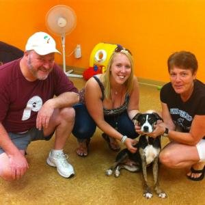 Three people and a dog sitting on the floor in a room with an orange wall, a fan, and a yellow box in the background.