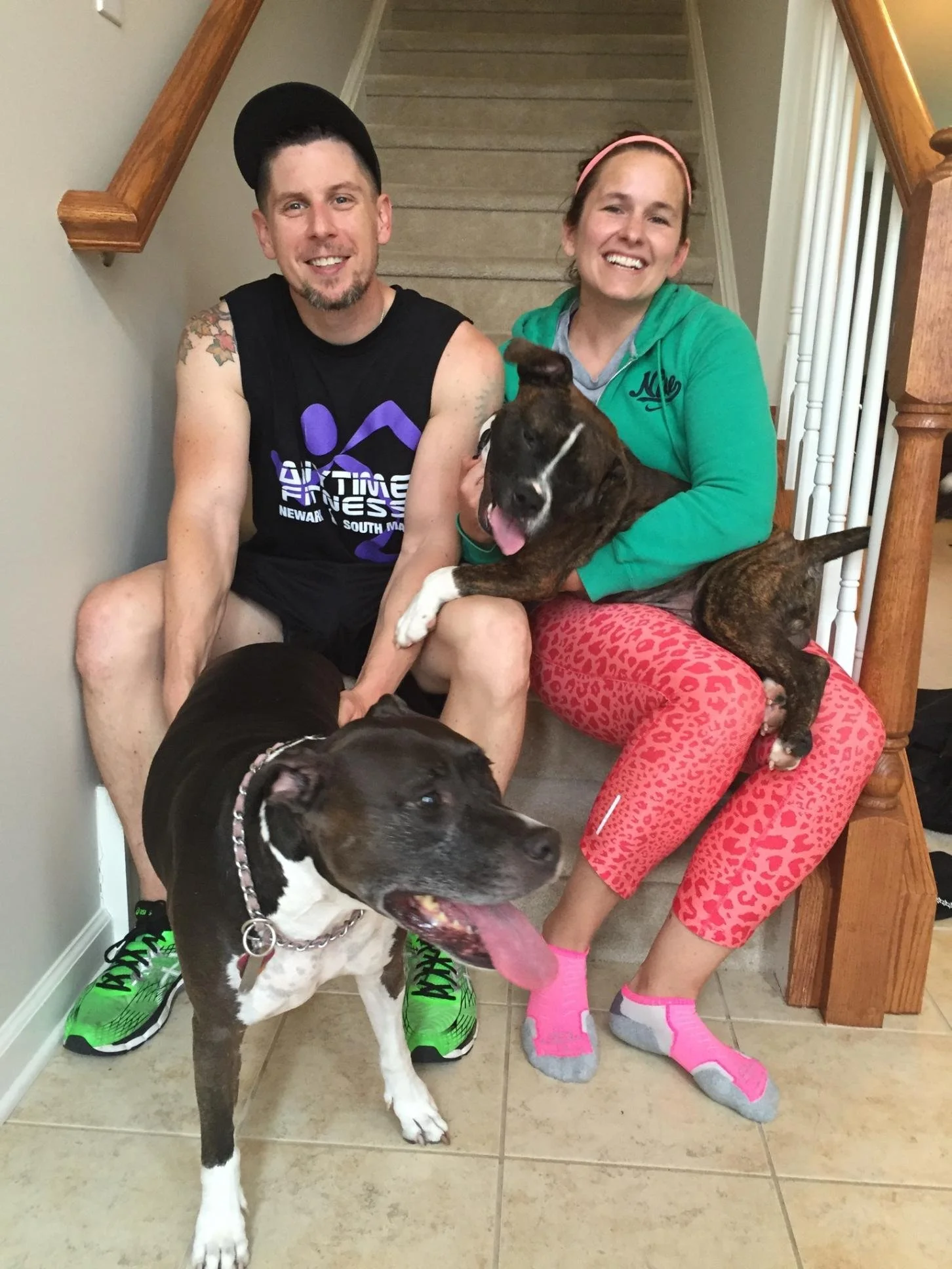 Two smiling women with two large dogs on a tiled staircase. One woman is wearing a black sleeveless shirt, black cap, and bright green sneakers. The other woman is wearing a green hoodie, pink patterned pants, and pink slippers. One dog is black and 