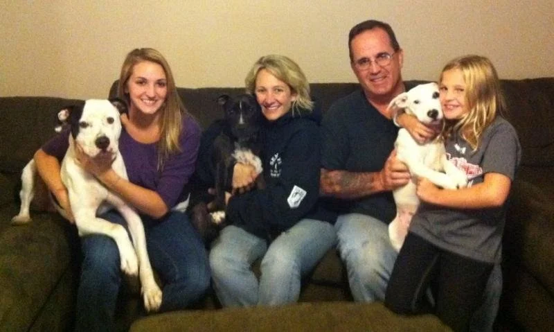 A family of five sitting on a couch, each holding or sitting next to a dog. The family includes a man, woman, two girls, and a woman with blonde hair. The dogs are two white with black spots, one on each side of the family.