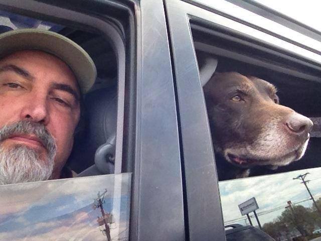 A man with a beard and gray cap taking a selfie with a dog in a vehicle, both looking out the window.