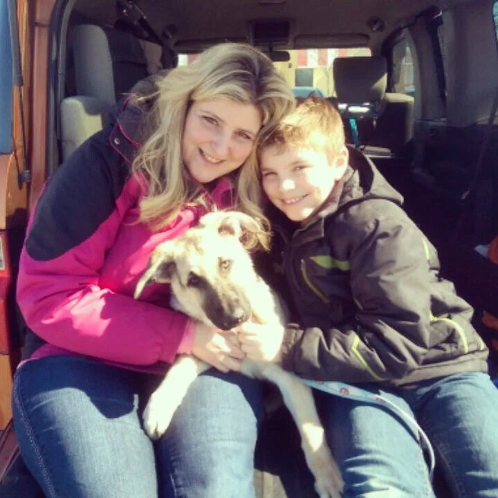 A woman and a boy sitting inside a vehicle, smiling at the camera, holding a puppy between them.