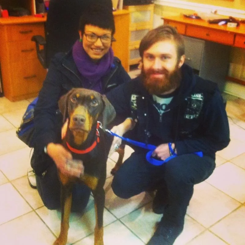 Two smiling people, one with glasses and a purple scarf and the other with a beard, are kneeling inside a room with tiled flooring, holding a brown dog with a red collar and blue leash.
