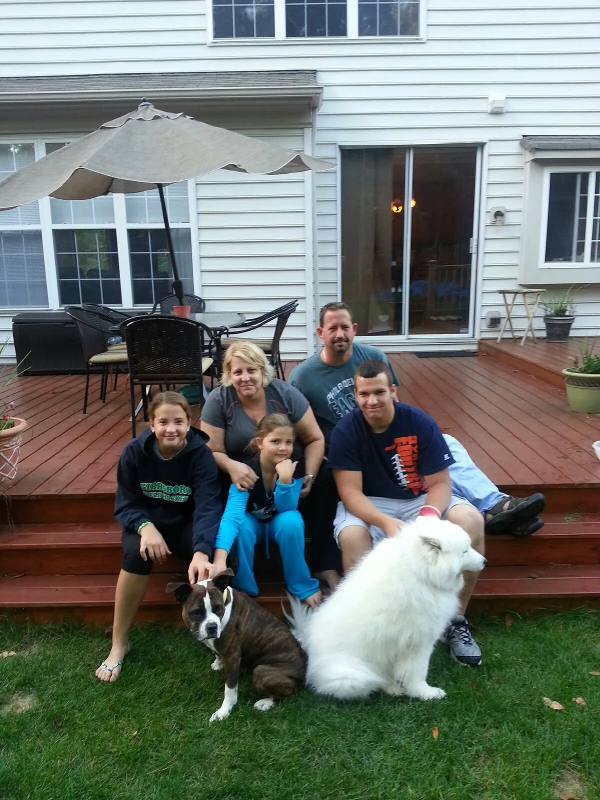 A family sitting on a wooden deck with two dogs, surrounded by potted plants, in front of a house with sliding glass door and windows.