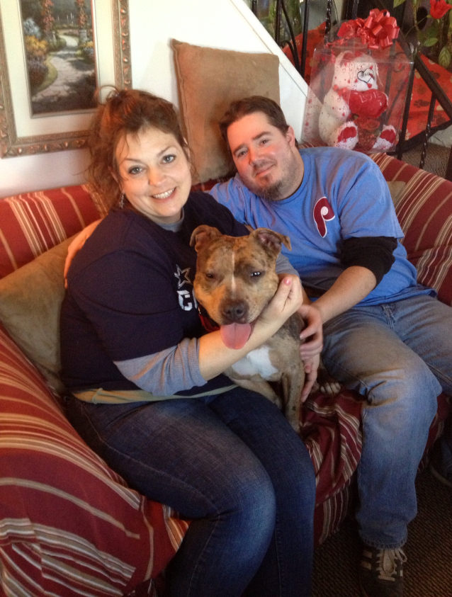 Two people sitting on a striped couch with a brown and white dog in front of them, indoors with a picture frame, flower arrangement, and Valentine's Day decorations behind.