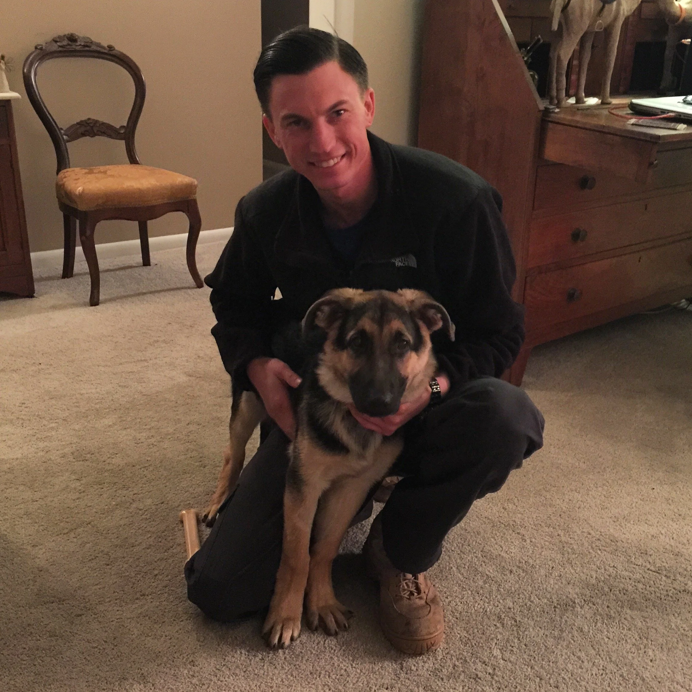 A young man with dark hair styled to the side, wearing a black North Face jacket, is sitting on the floor and holding a large, fluffy puppy with brown, black, and white fur. They are inside a room with carpeted flooring, wooden chairs, a dresser, and