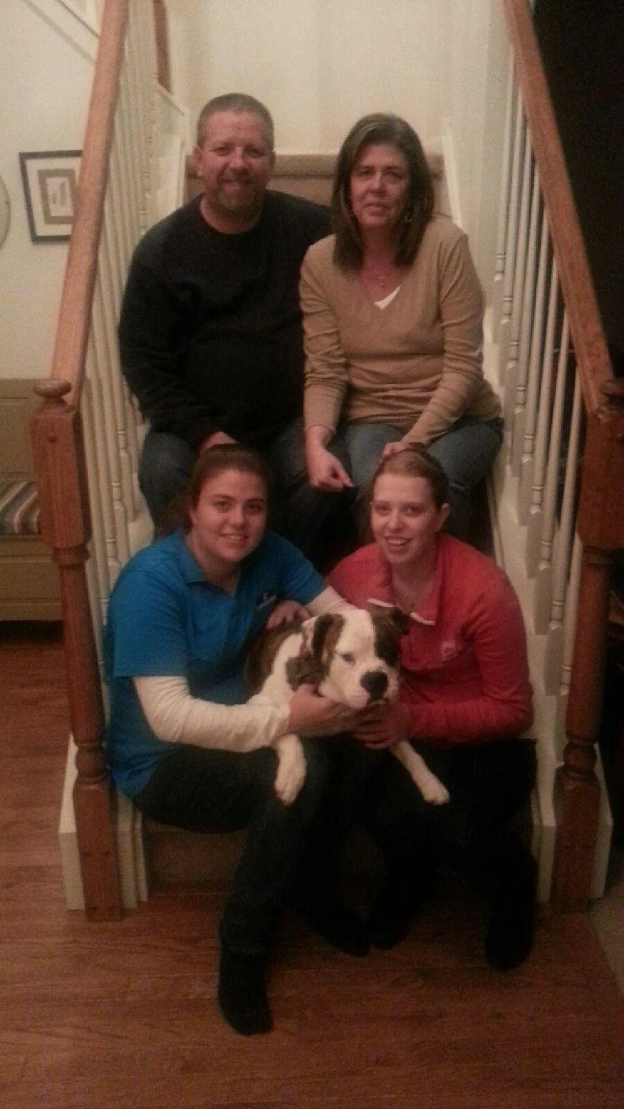 A family photo with five members, including a dog, sitting on a staircase in a home. Four people are visible: a man with a beard in a black shirt, a woman in a beige top, a young woman in a red shirt, and another young woman in a blue shirt holding a