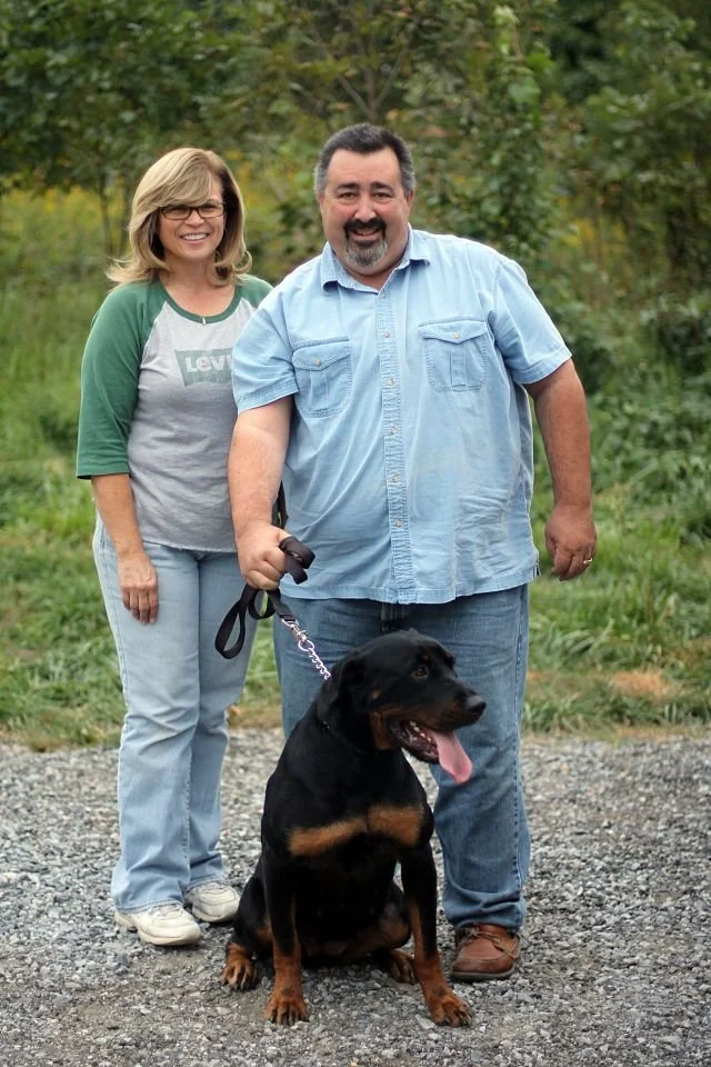 A smiling couple walking a Rottweiler dog outdoors on a gravel path with greenery in the background.