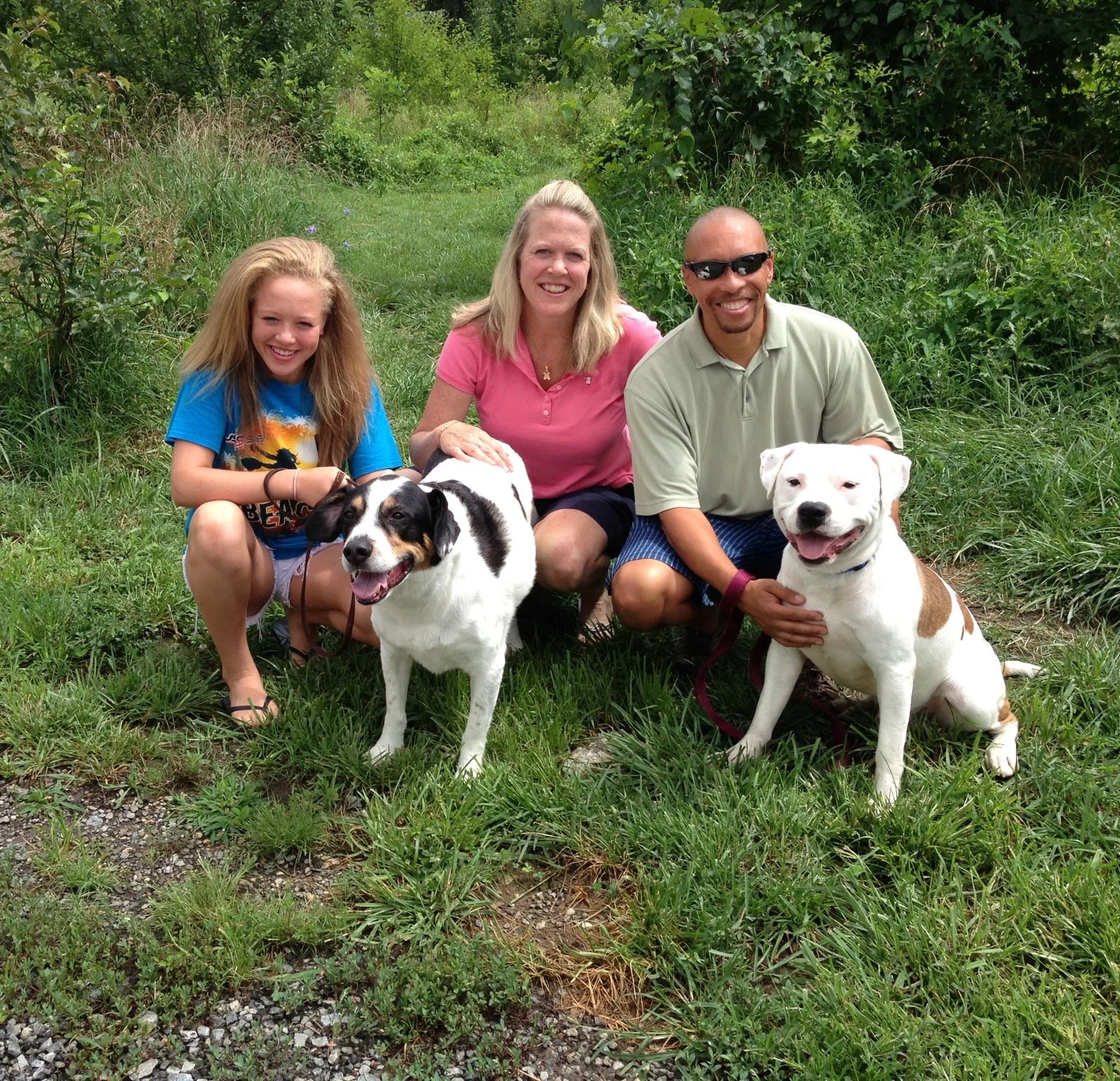 A group of three people and two dogs sitting on grass in a lush green outdoors setting. The group includes a young girl, a woman, and a man. The girl on the left has long blonde hair, is wearing a blue t-shirt, and is smiling. The woman in the middle