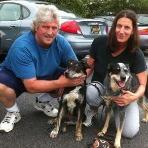 Two women crouching outdoors with two dogs, one black with white markings and the other black and white, in a parking lot with cars in the background.