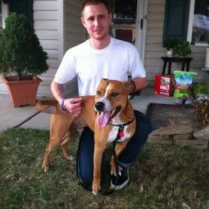 A man kneeling on a lawn with a brown and white dog, both facing the camera. The man is wearing a white T-shirt and dark pants. The background shows a house, potted plants, and a door.