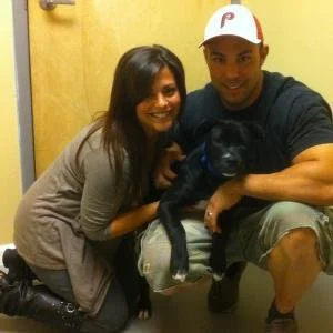 A man and a woman kneeling with a black puppy indoors, smiling at the camera.