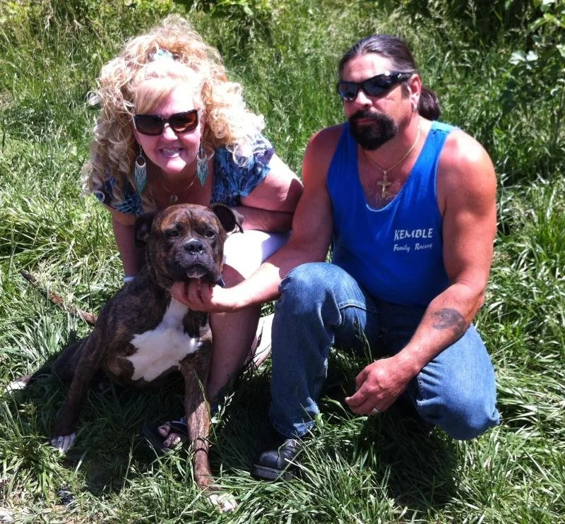 A woman and a man crouch outdoors on the grass with a brindle pitbull dog. The woman has curly blonde hair, sunglasses, and wears a patterned top. The man has long hair, a beard, sunglasses, and is wearing a blue sleeveless shirt and jeans. The woman