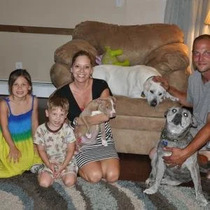 Family sitting on a living room floor with two small dogs and a pug sitting on a large beige couch.