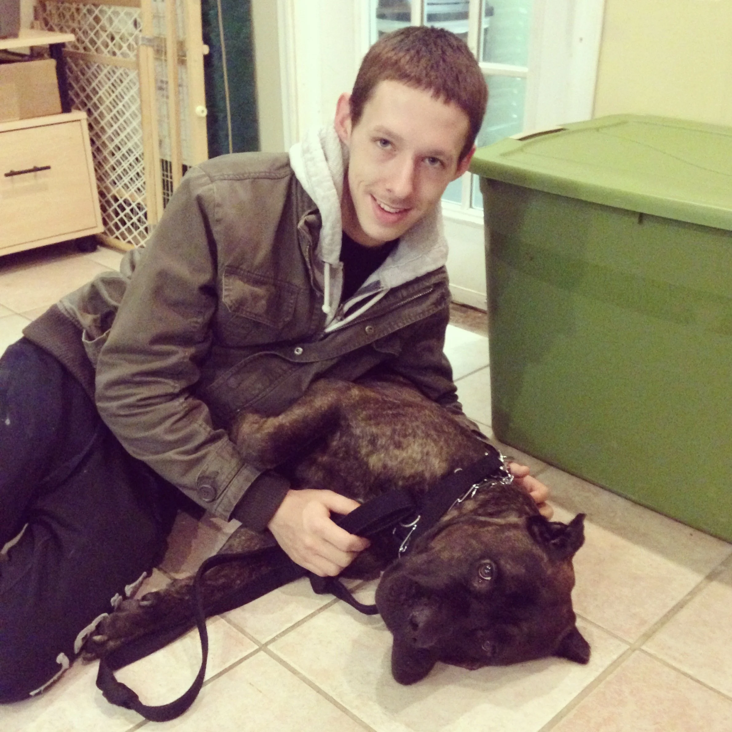 A young man with red hair and fair skin, wearing a brown jacket, is crouched on the floor indoors near a large green trash bin. He is smiling and looking at the camera, with a black dog in front of him. The background shows a closed sliding glass doo