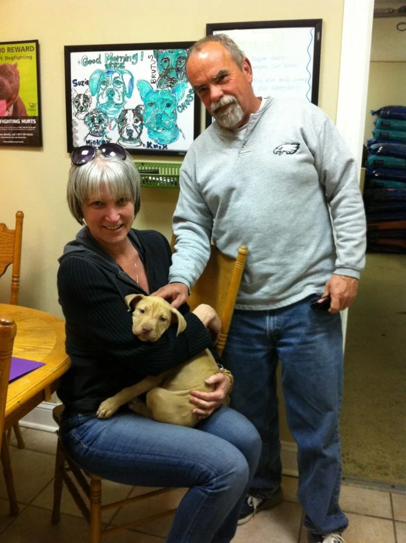 A woman sitting on a wooden chair holding a tan puppy, with an older man standing next to her in an indoor setting. The woman has short gray hair and sunglasses on her head. The man has gray hair and a beard, wearing a light gray sweatshirt with an e