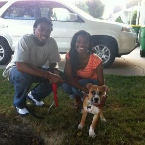 A man and a woman kneeling on the grass, holding a leash attached to a dog, with a white SUV in the background.