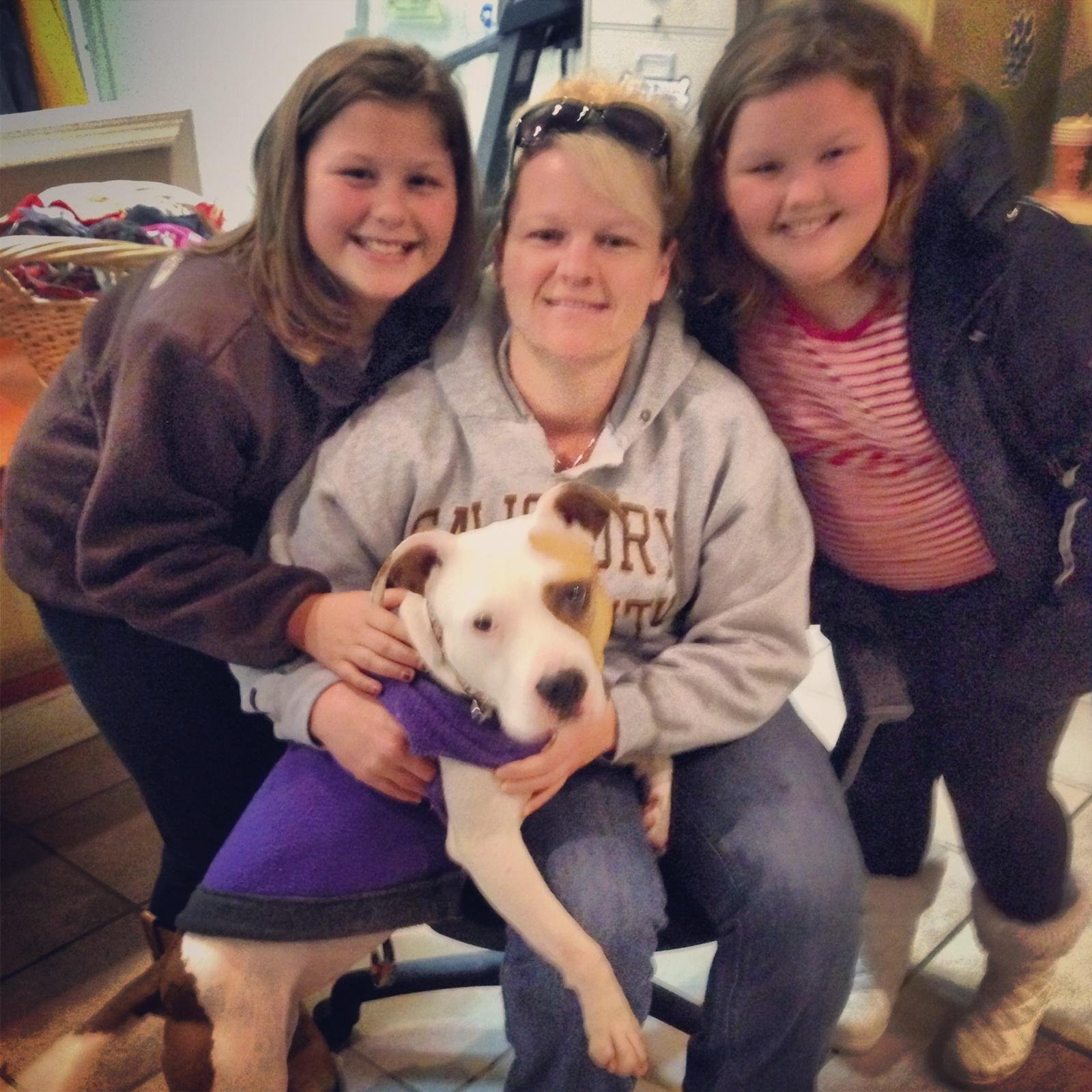 Three women and a puppy sitting on a bench in a cafe. The woman in the middle is holding the puppy, which is wearing a purple sweater. The women are smiling and posing for the photo.