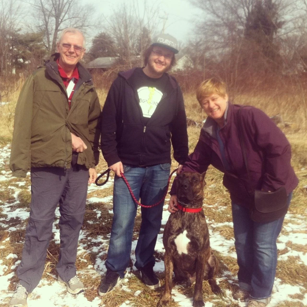 Three people standing outdoors in a field with snow patches, smiling with a brindle-colored dog sitting between them. The person in the middle is holding the dog's leash.