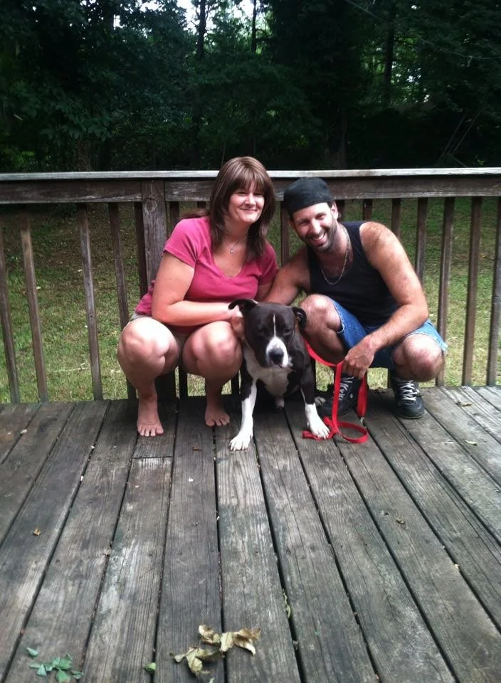 A woman and a man squatting on a wooden deck with a black and white dog between them, surrounded by trees.