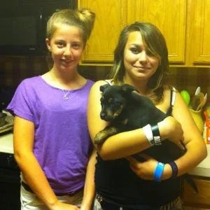 Two girls standing in a kitchen, one holding a black puppy.