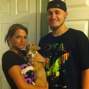 A woman and a man smiling and holding a small brown and white puppy