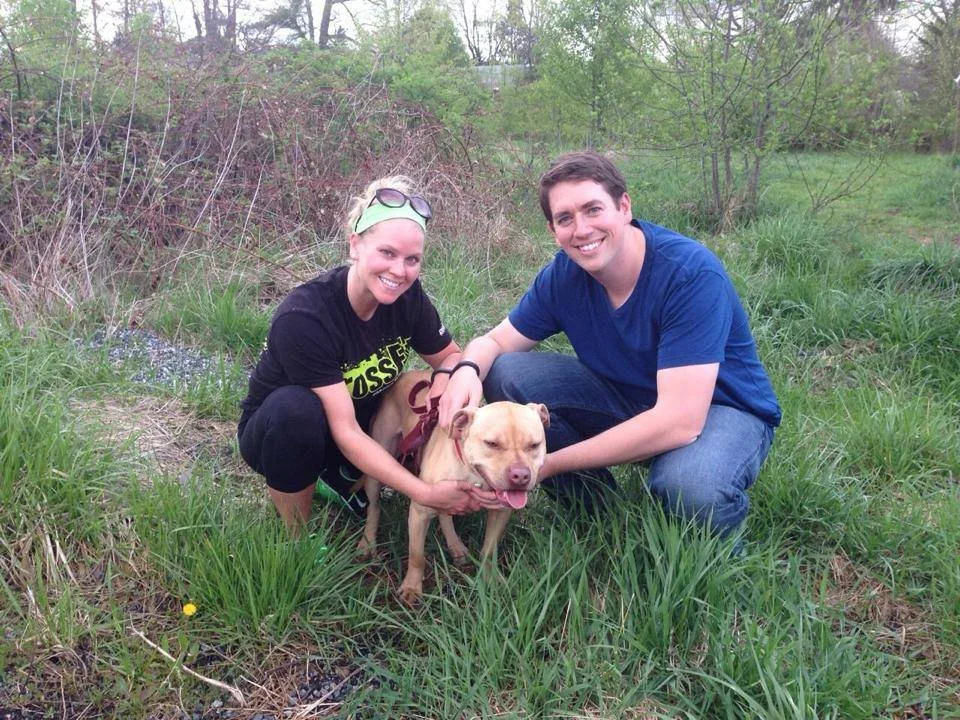 A smiling couple with a dog in a grassy outdoor area surrounded by trees and bushes.