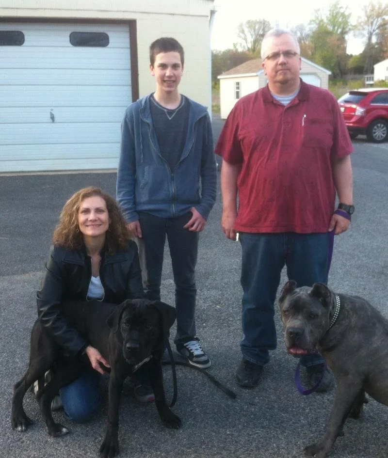 A woman kneeling next to a black dog, a young man standing, and a man standing with a gray dog, in a driveway with a garage and houses in the background.
