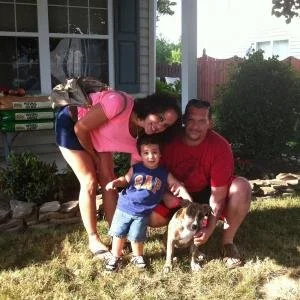Family of three with a dog posing on a lawn in front of a house, smiling and enjoying the outdoors.