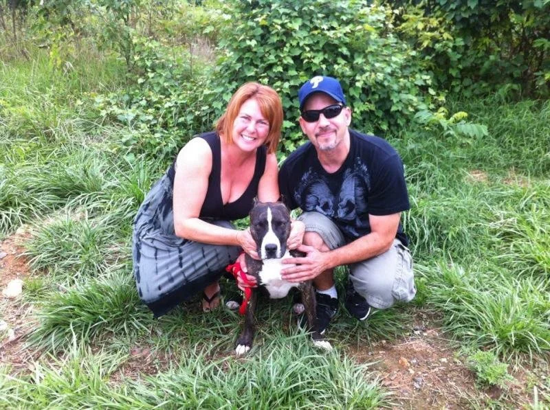 A woman and a man kneeling on grass, smiling, with their hands on a black and white dog wearing a red collar, outdoors surrounded by greenery.