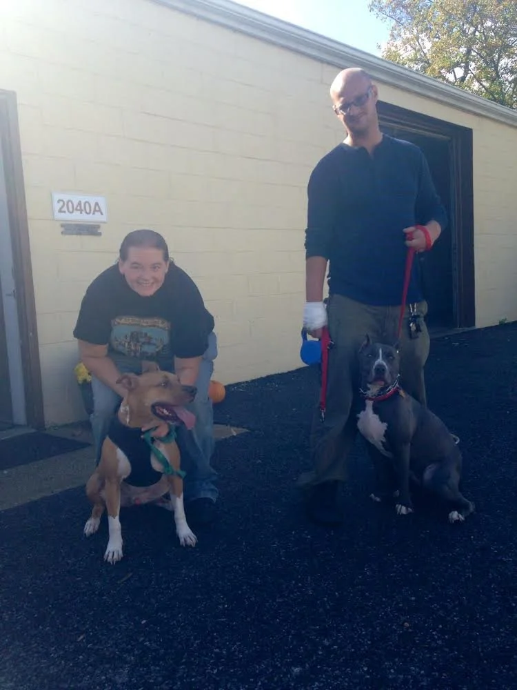 Two people with their dogs outside a light-colored building, one boy crouching with a brown and white dog, and a man standing with a black and white dog on a leash.