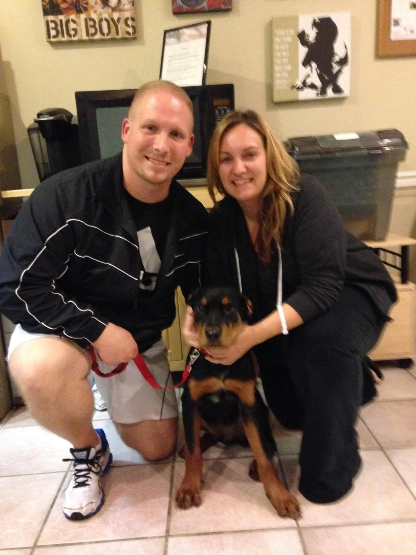 A man and woman pose with a Rottweiler dog in a room decorated with war art and posters on the wall.