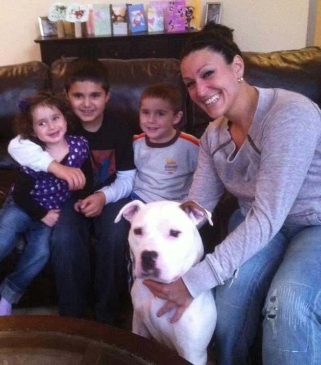 A woman and four children with a white pit bull puppy sitting on her lap, all smiling and sitting on a brown couch in a living room, with greeting cards and pictures on a shelf in the background.