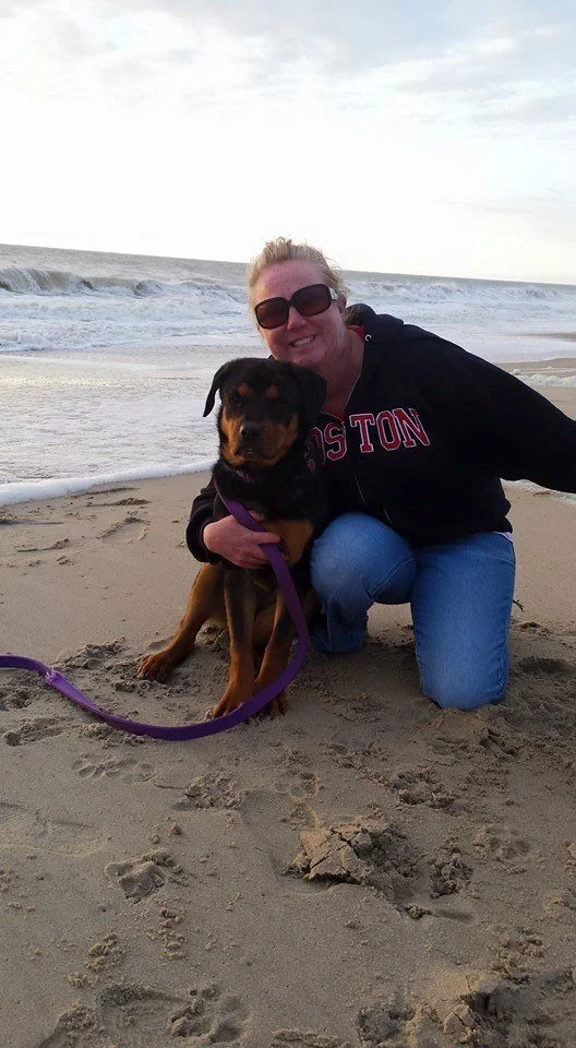 A woman with blonde hair, wearing sunglasses and a black hoodie with red lettering, is smiling and kneeling on a sandy beach with her arm around a black and brown Rottweiler puppy on a purple leash. The ocean waves are in the background.