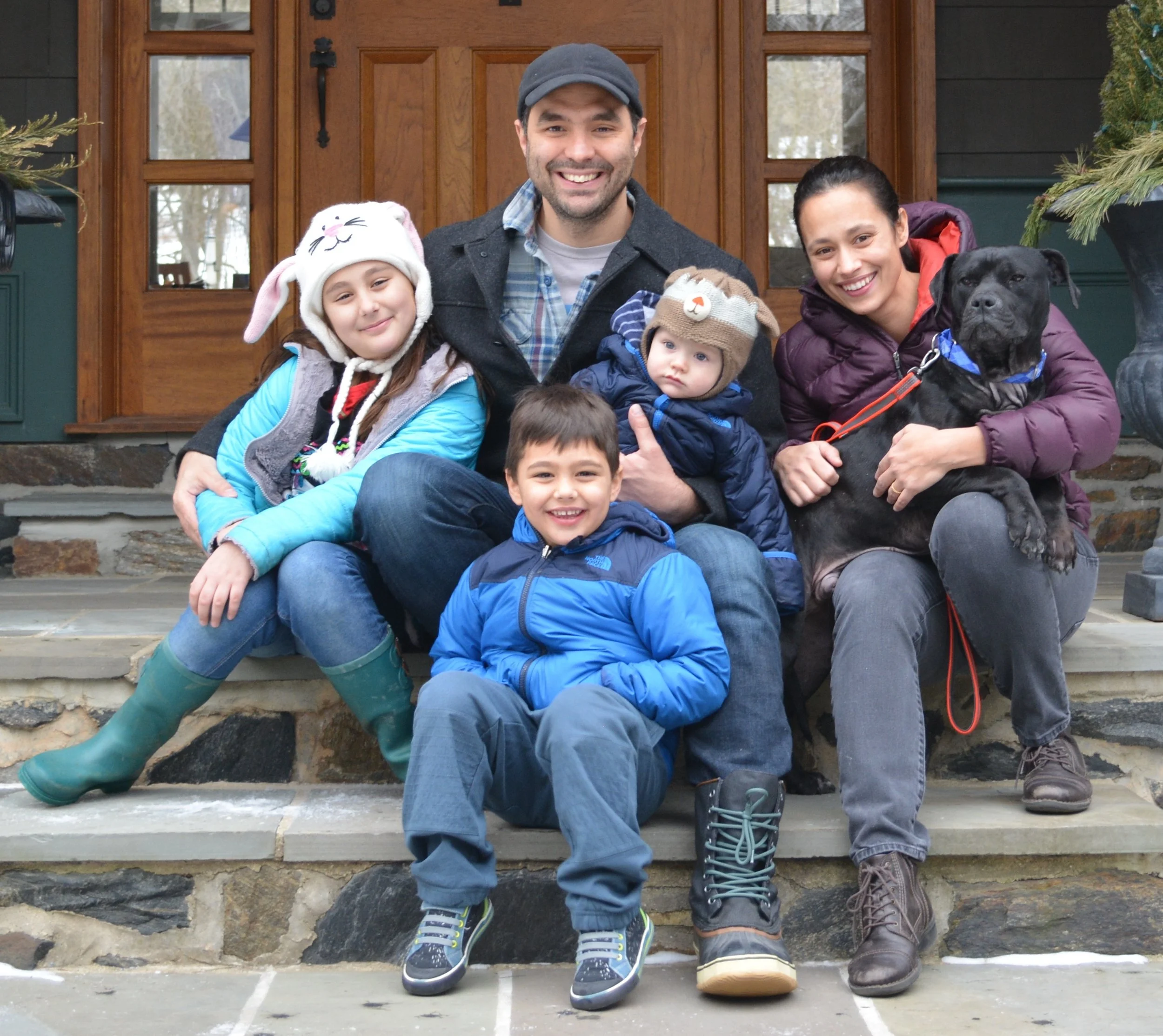 A smiling family of five and their dog sitting on front steps of a house. The children are wearing winter clothing, one with a bunny hat, and the dog is a black Labrador with a blue bandana. The adult woman has dark hair pulled back, and the man is w